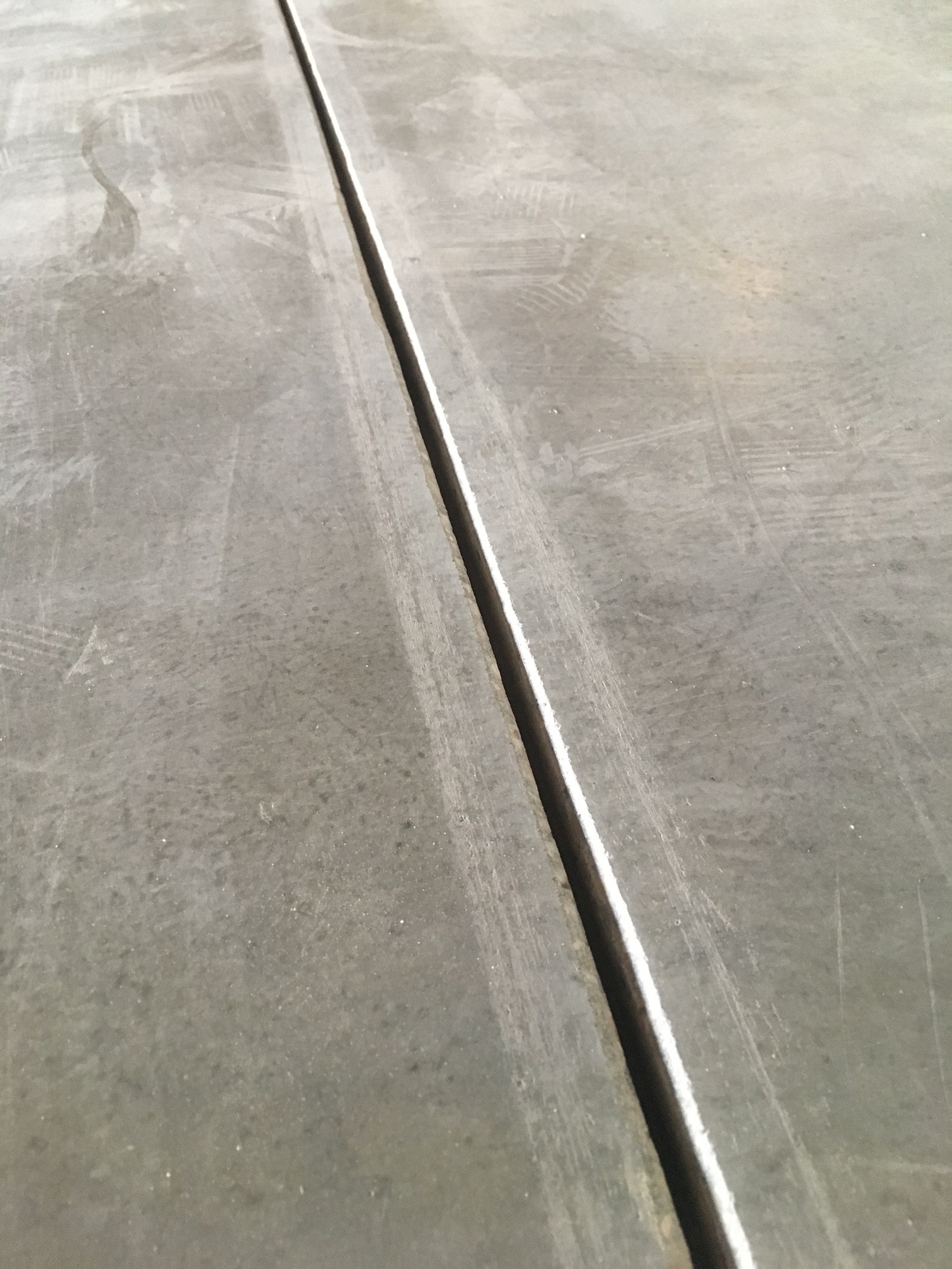 Szara, gładka powierzchnia z wyraźnym dylatacyjnym nacięciem pośrodku. Widoczne ślady po narzędziach i delikatne zarysowania na powierzchni. Minimalistyczne ujęcie.