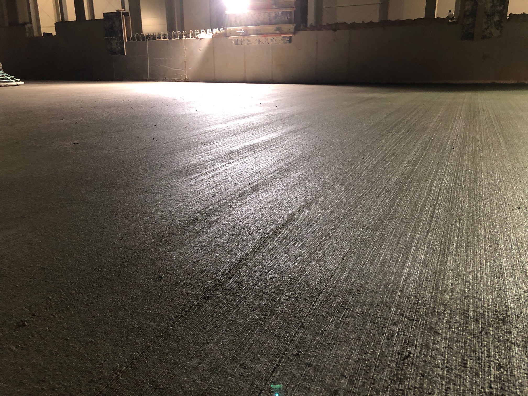 Świeżo wylana, szara posadzka z widocznymi śladami po zacieraniu, oświetlona mocnym światłem. W tle fragment ściany i konstrukcji budynku w surowym stanie.