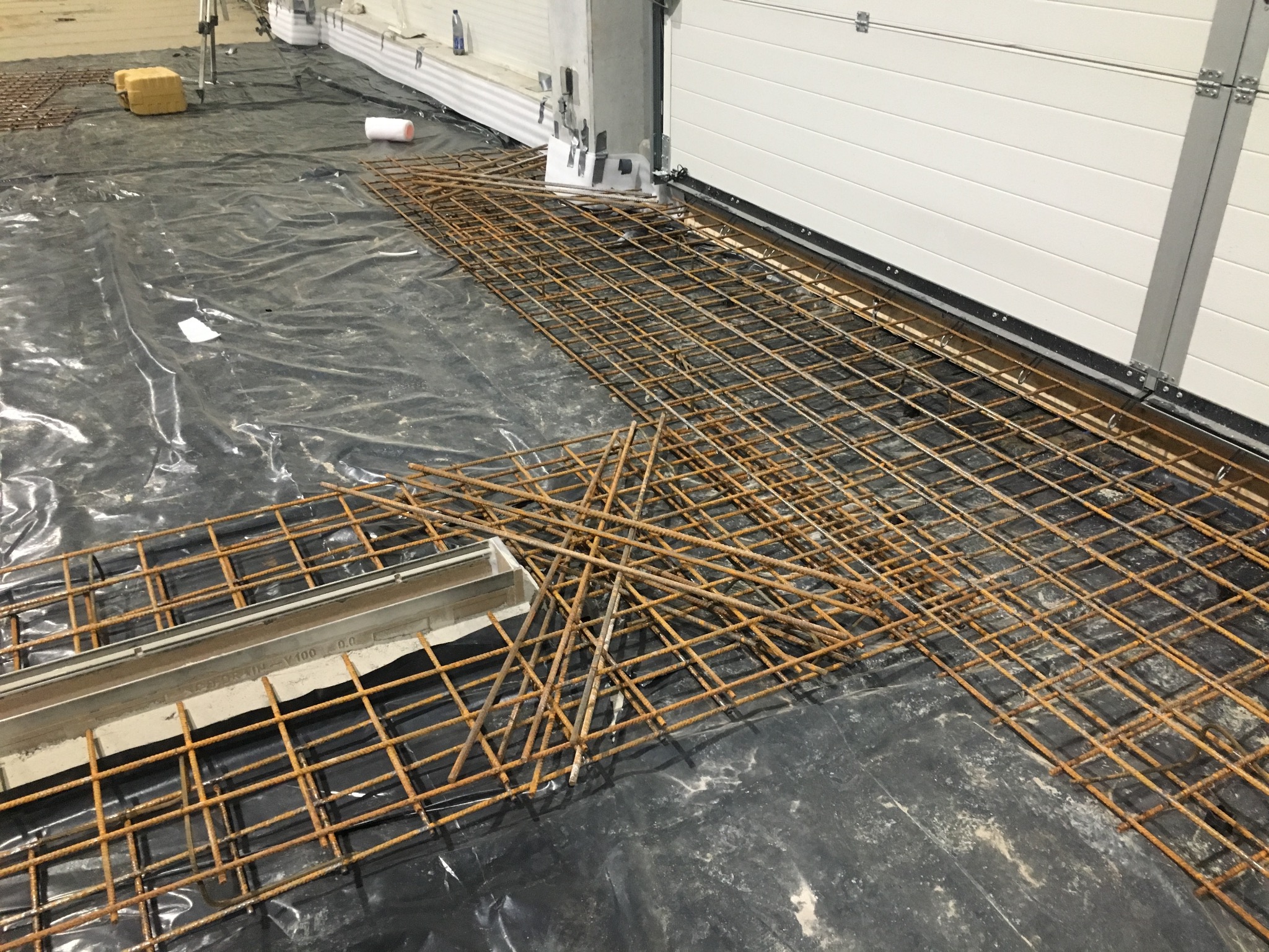 Przygotowanie do wylewki: rozłożona siatka zbrojeniowa na folii, widoczne elementy szalunku przy bramie garażowej. Etap prac przed zalaniem betonem.