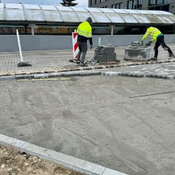 SURIS SP. Z O.O. - Dwóch pracowników w odblaskowych kamizelkach układa kostkę brukową o heksagonalnym kształcie na przygotowanym podłożu z piasku i cementu, widoczne ułożone obrzeża, paleta z kostką i butelka wody...