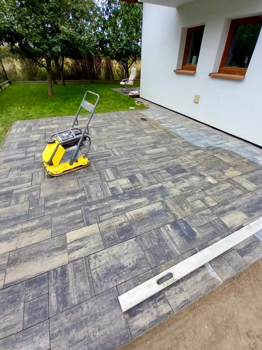 Fragment układanej nawierzchni z kostki brukowej w odcieniach szarości i beżu, z żółtą zagęszczarką wibracyjną i poziomnicą aluminiową, widok z góry.