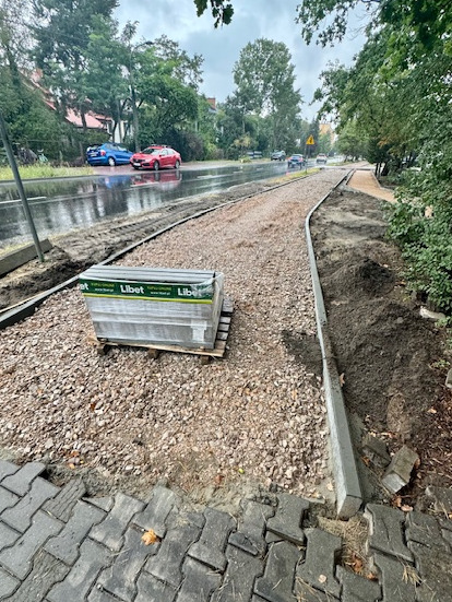 Przygotowanie podłoża pod chodnik: warstwa żwiru, obrzeża betonowe i paleta kostki brukowej Libet na wilgotnej nawierzchni ulicy w deszczowy dzień.