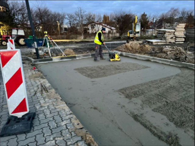 Pracownik w kamizelce odblaskowej zagęszcza wibro-płytą świeżo wylany beton na podbudowie pod chodnik z kostki brukowej, widoczny znak ostrzegawczy, w tle sprzęt budowlany.