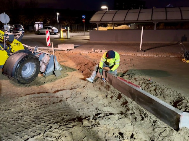 Pracownik w odblaskowej kamizelce układa obrzeża chodnika z kostki brukowej, wspierając się o kolano, w tle widoczny żółty mini-ładowarka i ubijak wibracyjny.