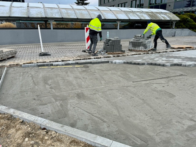 Dwóch pracowników w odblaskowych kamizelkach układa kostkę brukową o heksagonalnym kształcie na przygotowanym podłożu z piasku i cementu, widoczne ułożone obrzeża, paleta z kostką i butelka wody...