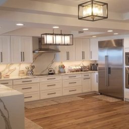 KOSSAKO - Nowoczesna, jasna kuchnia z wyspą i marmurowym blatem. Białe szafki z czarnymi uchwytami, drewniana podłoga, designerskie lampy. Widok na całe pomieszczenie.