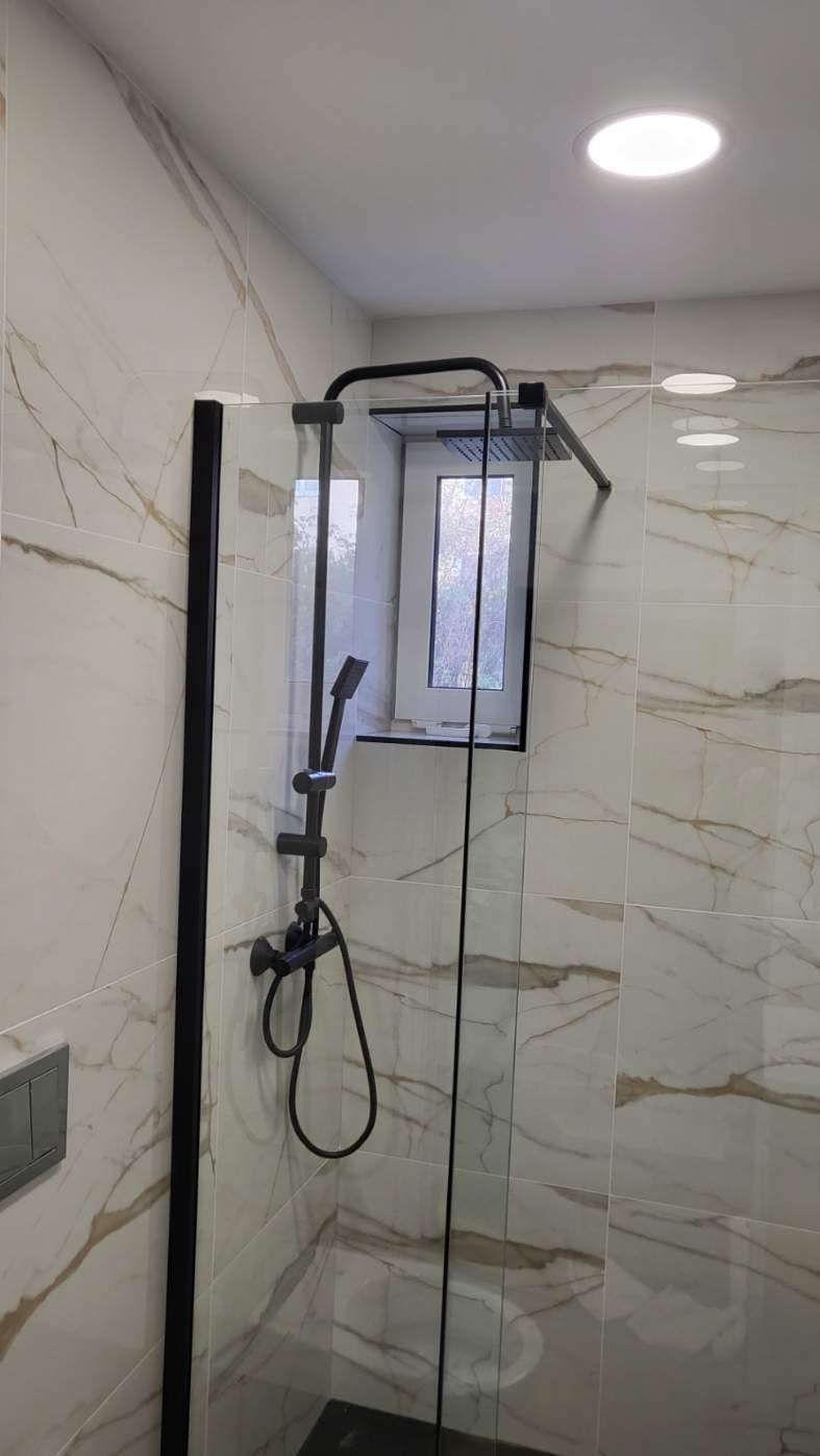 Nowoczesna kabina prysznicowa z czarną armaturą i marmurowymi płytkami w wyremontowanej łazience. Okno nad kabiną. Oświetlenie sufitowe.