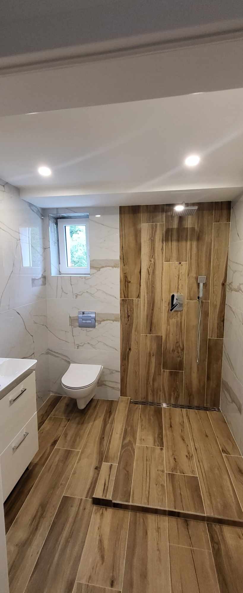 Wykończona łazienka z drewnopodobnymi płytkami na podłodze i ścianie prysznicowej, marmurowe płytki na pozostałych ścianach, biała armatura i szafka pod umywalką.