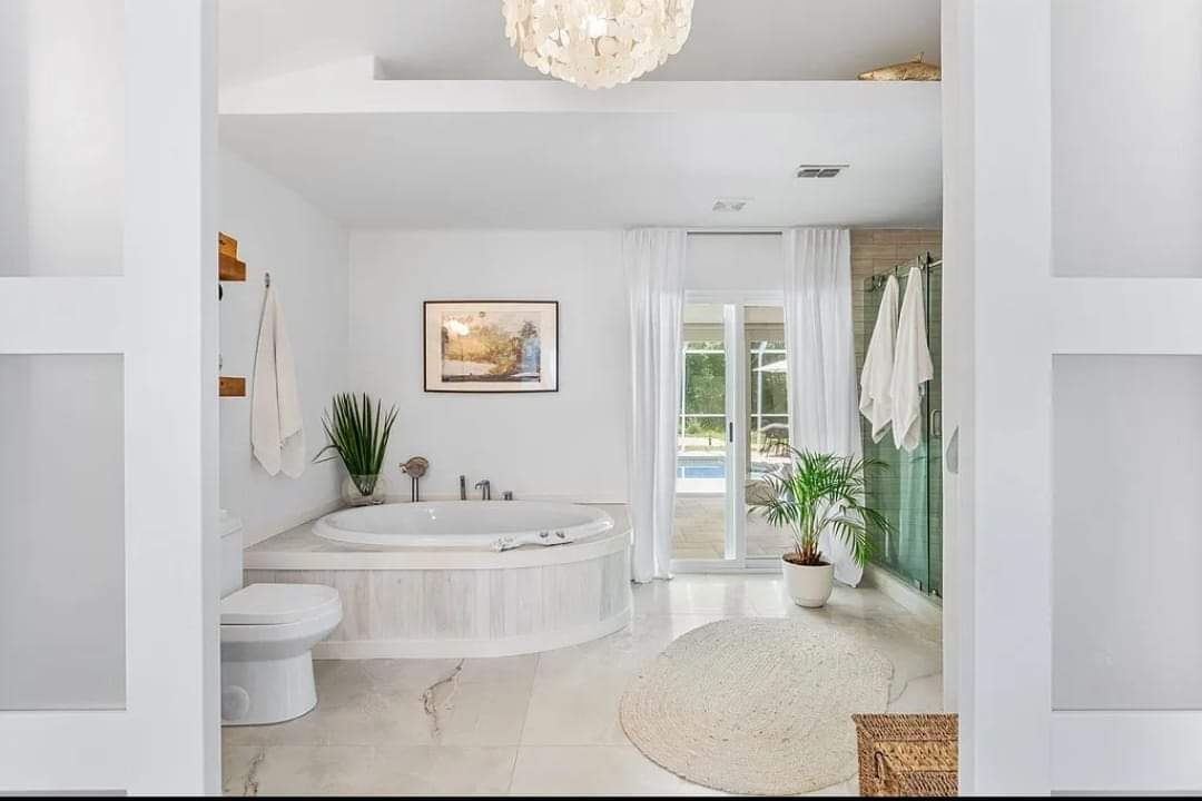 Jasna łazienka z okrągłą wanną obudowaną drewnem, marmurową podłogą i roślinami. Widok na basen przez przesuwne drzwi. Lampa w stylu muszli.