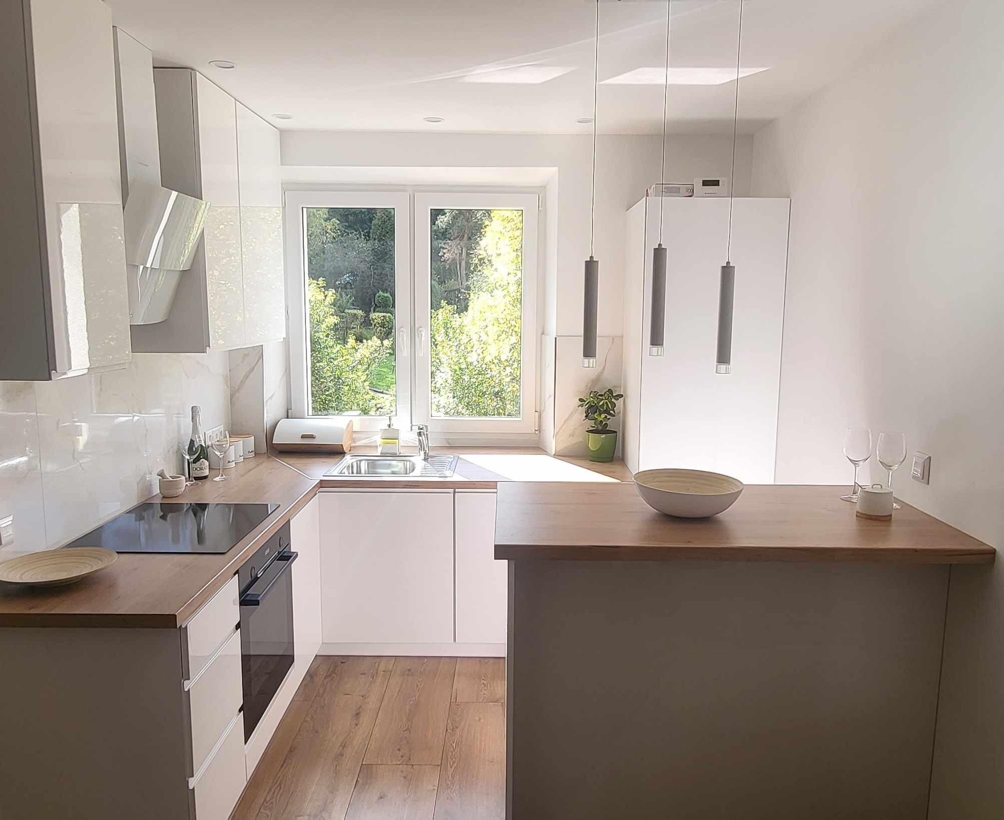 Jasna kuchnia z drewnianym blatem i widokiem na zieleń za oknem. Minimalistyczne lampy wiszące, białe szafki i sprzęty AGD tworzą nowoczesny wystrój.