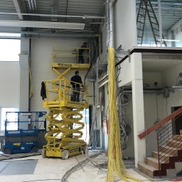 NAFIIS SPÓŁKA Z OGRANICZONĄ ODPOWIEDZIALNOŚCIĄ - Elektrycy na podnośnikach montują kable w nowym budynku, widoczne wiązki żółtych kabli, niebieski podnośnik i metalowe koryta kablowe pod sufitem.