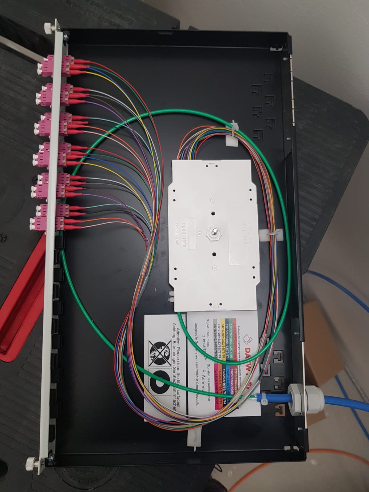 Panel krosowniczy światłowodowy z podłączonymi kablami w różnych kolorach, widoczne złącza światłowodowe i etykieta z informacjami technicznymi.