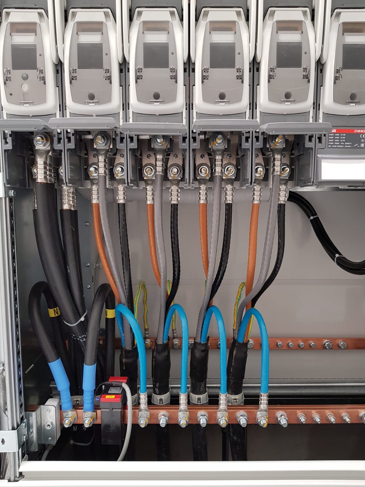 Wnętrze rozdzielnicy elektrycznej z widocznymi przewodami o różnych kolorach (czarny, szary, pomarańczowy, niebieski) podłączonymi do listew zaciskowych, z zamontowanymi licznikami i modułem...