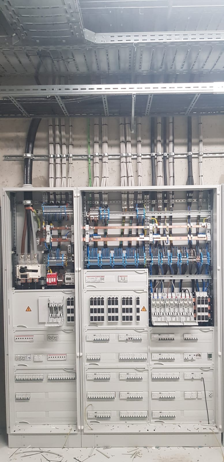 Rozdzielnica elektryczna z otwartymi drzwiami, widoczne liczne kable, bezpieczniki i elementy sterujące, pod sufitem metalowe korytka kablowe z poprowadzonymi przewodami.