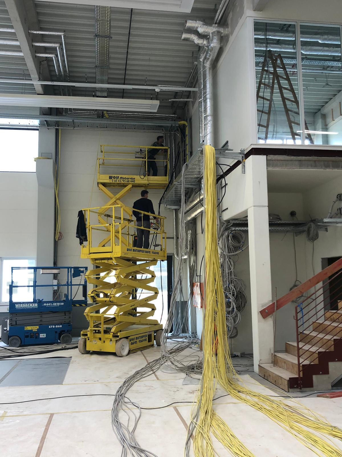 Elektrycy na podnośnikach montują kable w nowym budynku, widoczne wiązki żółtych kabli, niebieski podnośnik i metalowe koryta kablowe pod sufitem.