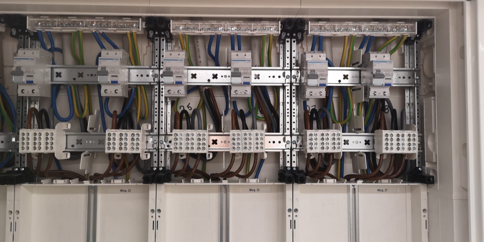 Rozdzielnica elektryczna w obudowie, widoczne zabezpieczenia, listwy zaciskowe i przewody w kolorach niebieskim, żółto-zielonym i brązowym, zamontowane na szynach DIN.