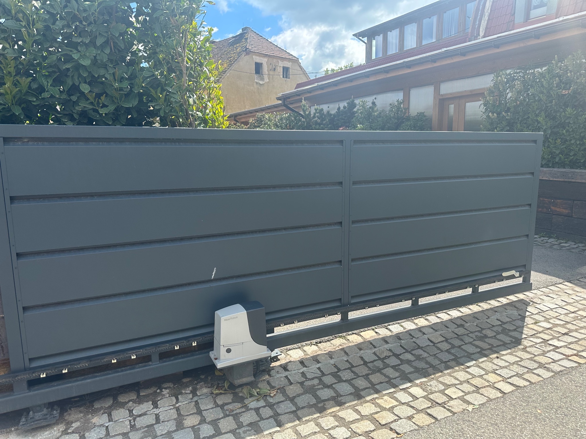 Nowoczesna, przesuwna brama w kolorze antracytu z widocznym napędem Nice Robus na tle brukowanej nawierzchni i zieleni. Estetyczne wykończenie posesji.