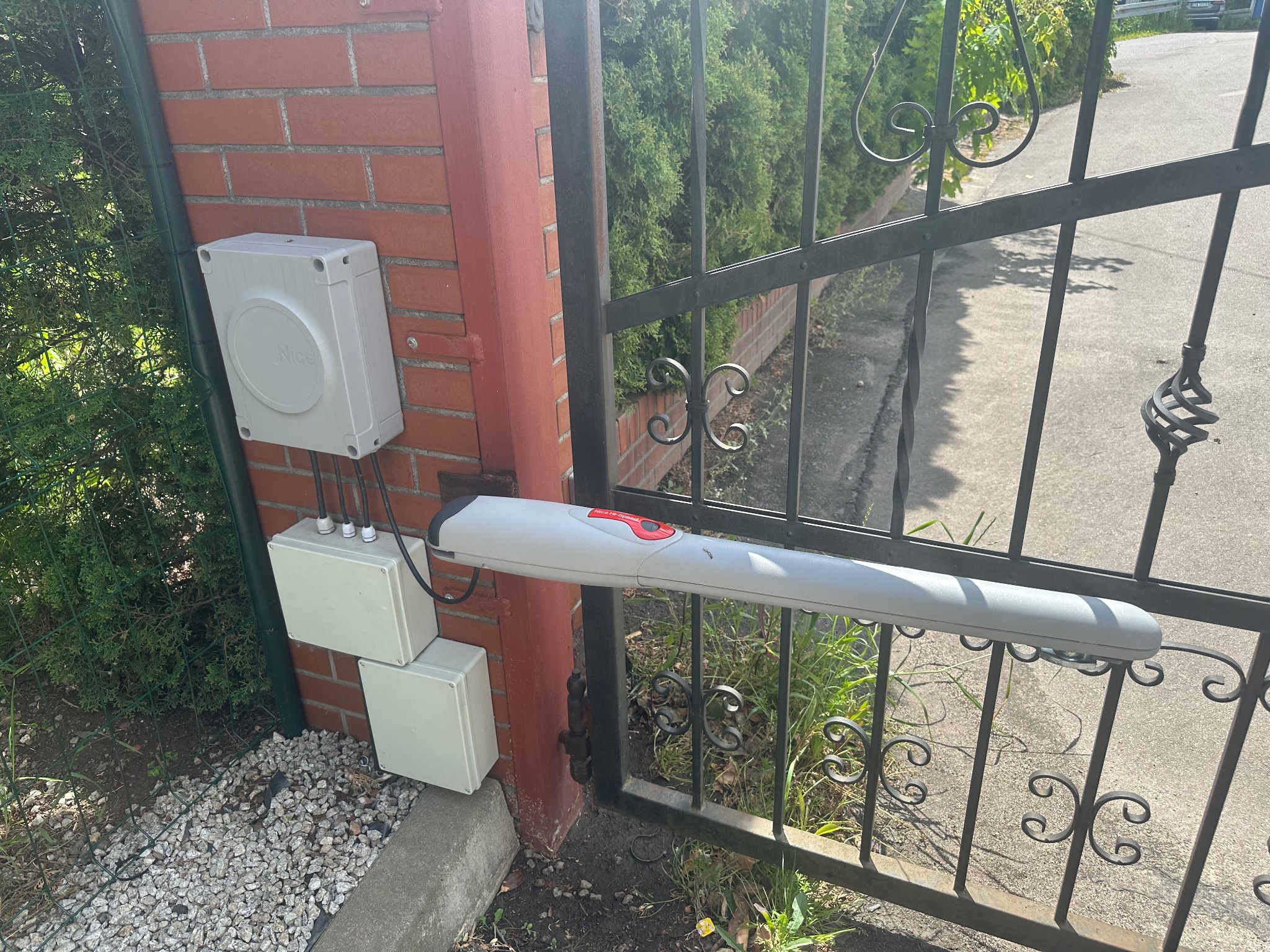 Zamontowany napęd bramy Nice z centralą sterującą na ceglanym słupku. Widoczne przewody i ozdobne elementy metalowej bramy. Tło z zielenią.