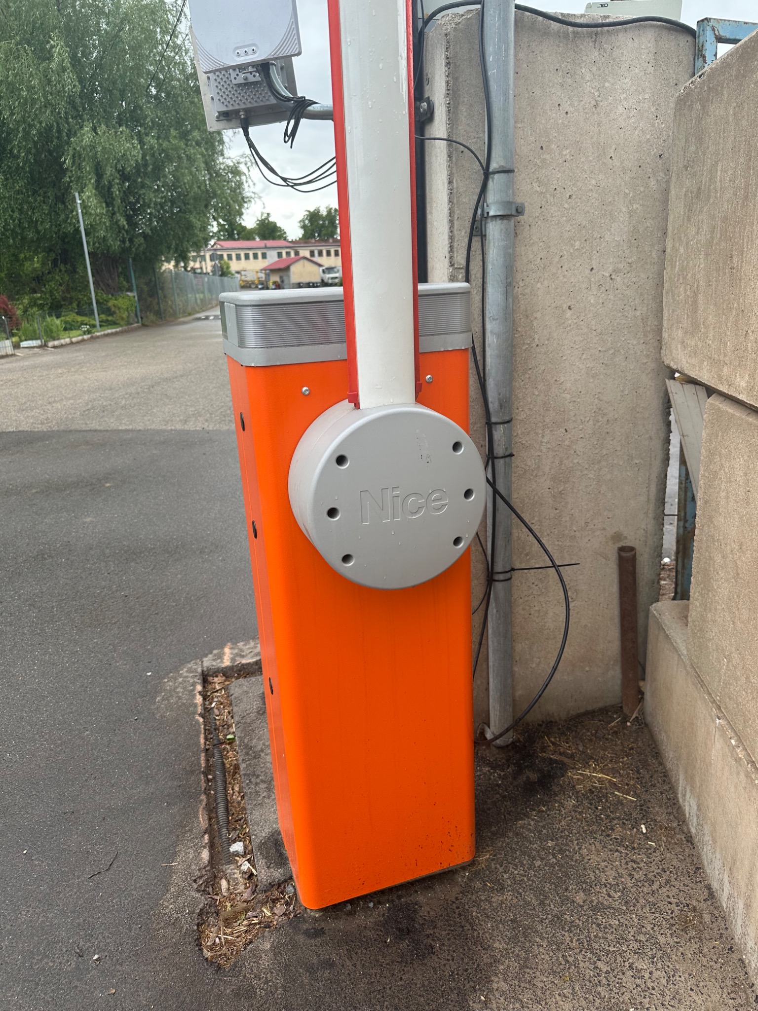 Pomarańczowy automat bramowy Nice z białą barierą, widoczny z boku na tle betonowego muru i asfaltowej drogi. Na automacie zamontowana antena.