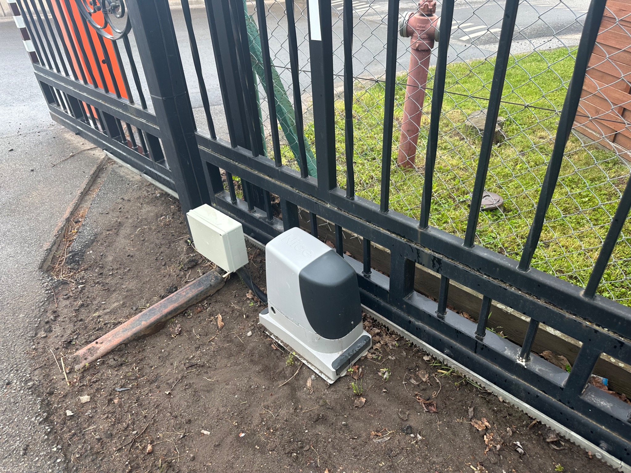 Szczegółowe ujęcie napędu bramy przesuwnej Nice, zamontowanego przy czarnej, metalowej bramie z pomarańczowymi elementami, widok na trawnik i hydrant.