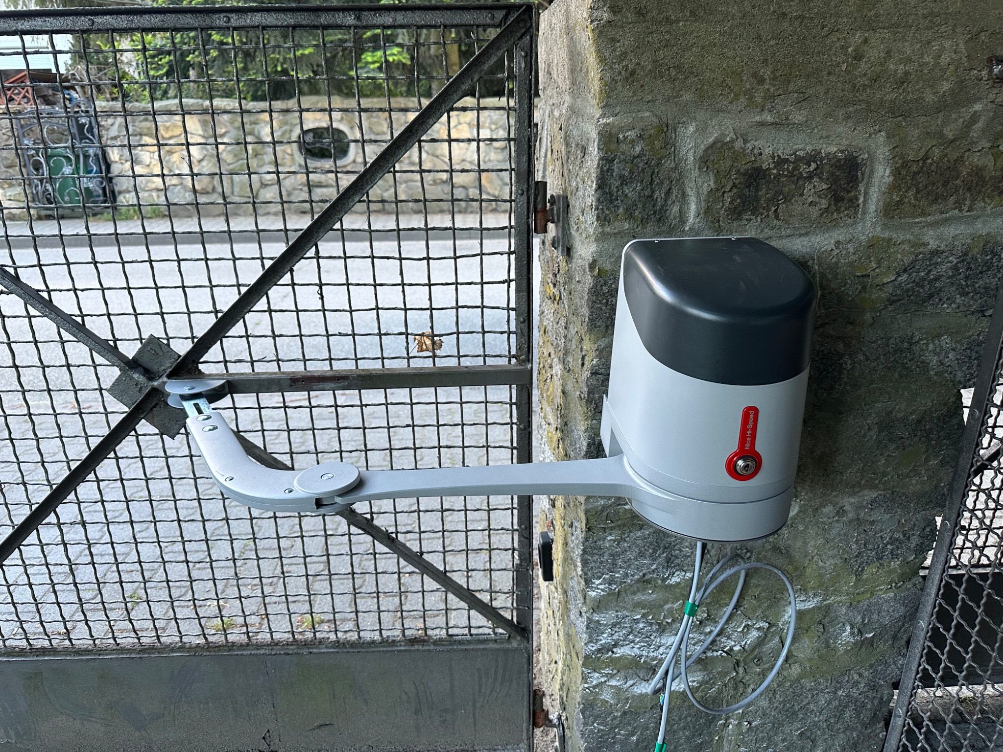 Zamontowany napęd Nice Hi-Speed do metalowej bramy skrzydłowej, widoczny na tle kamiennego słupka i metalowej siatki ogrodzenia.