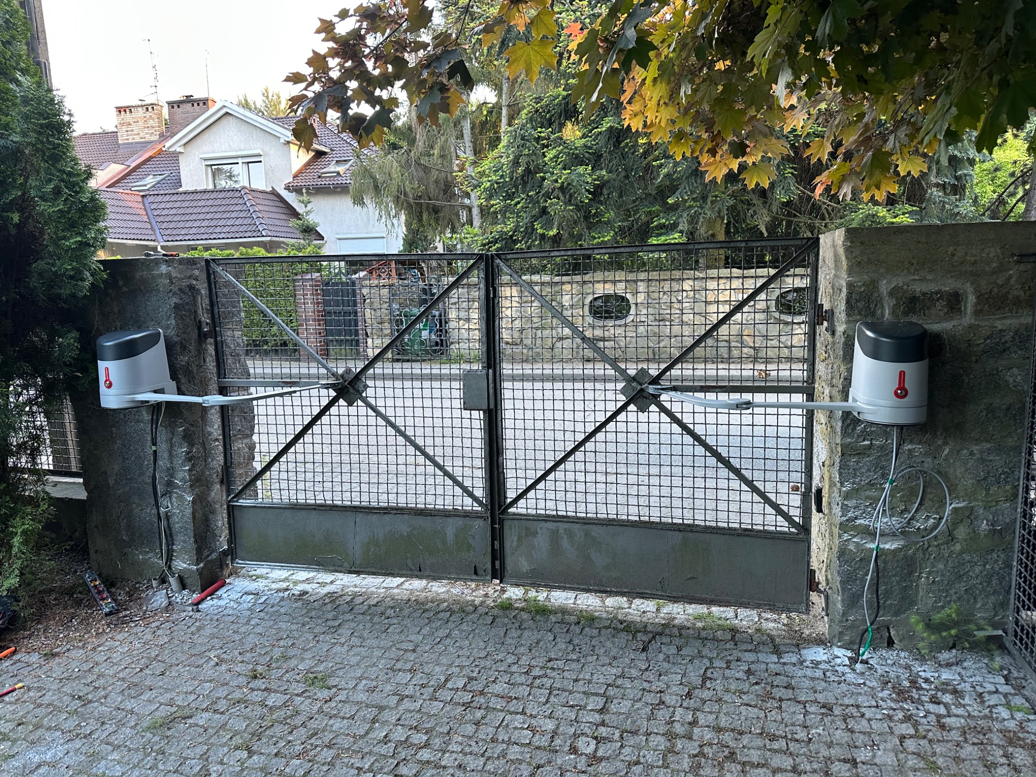 Automatyczna brama wjazdowa z napędem na kamiennych słupkach, widok z zewnątrz. Widoczne przewody i narzędzia sugerujące prace instalacyjne.