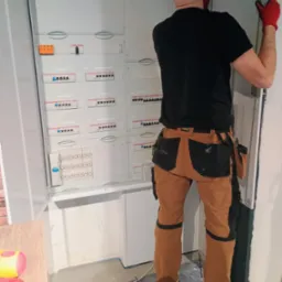 Elektryk w czerwonych rękawicach montuje rozdzielnicę elektryczną, widoczne zabezpieczenia i okablowanie wewnątrz szafy, narzędzia przy pasie, w tle fragment ściany z cegły.