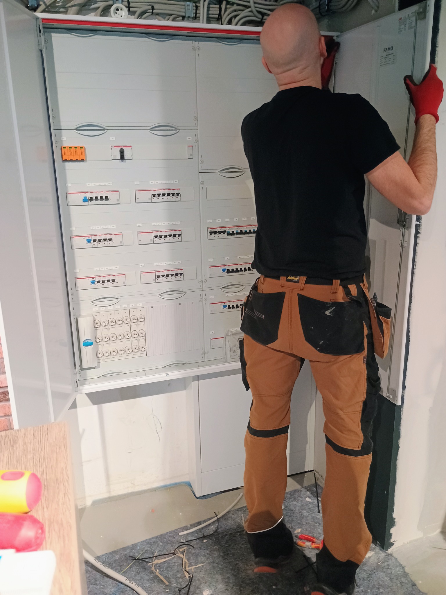 Elektryk w czerwonych rękawicach montuje rozdzielnicę elektryczną, widoczne zabezpieczenia i okablowanie wewnątrz szafy, narzędzia przy pasie, w tle fragment ściany z cegły.
