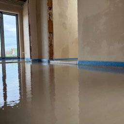 KN24 SPÓŁKA Z OGRANICZONĄ ODPOWIEDZIALNOŚCIĄ - Świeżo wylana, gładka posadzka w jasnym pomieszczeniu z oknem, zabezpieczona niebieską taśmą przy ścianach.