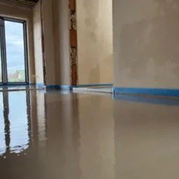 Świeżo wylana, gładka posadzka w jasnym pomieszczeniu z oknem, zabezpieczona niebieską taśmą przy ścianach.