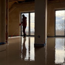 KN24 SPÓŁKA Z OGRANICZONĄ ODPOWIEDZIALNOŚCIĄ - Aplikacja wylewki samopoziomującej w jasnym pomieszczeniu z dużymi oknami, pracownik rozprowadza masę za pomocą rakli na długim kiju, zabezpieczone ściany taśmą malarską.