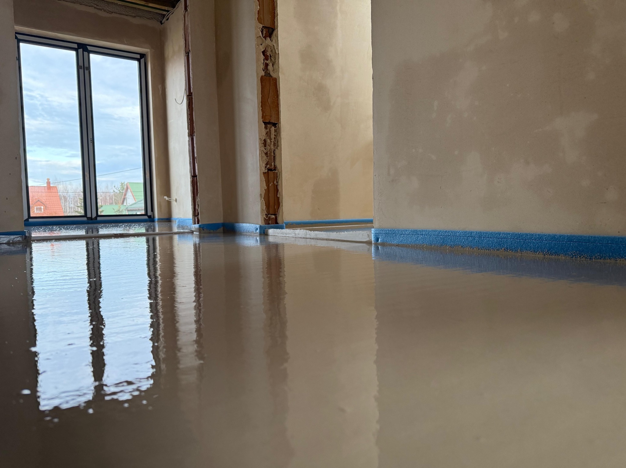 Świeżo wylana, gładka posadzka w jasnym pomieszczeniu z oknem, zabezpieczona niebieską taśmą przy ścianach.