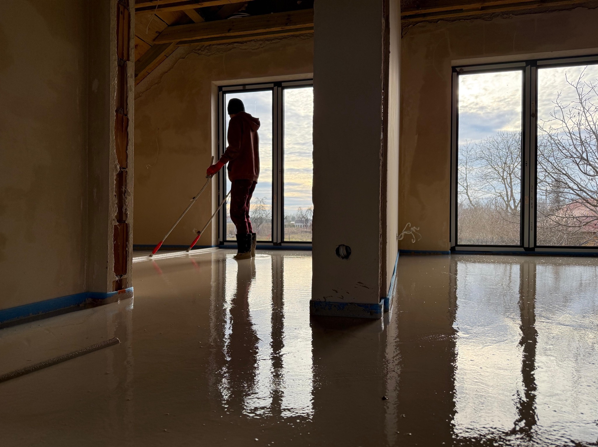 Aplikacja wylewki samopoziomującej w jasnym pomieszczeniu z dużymi oknami, pracownik rozprowadza masę za pomocą rakli na długim kiju, zabezpieczone ściany taśmą malarską.