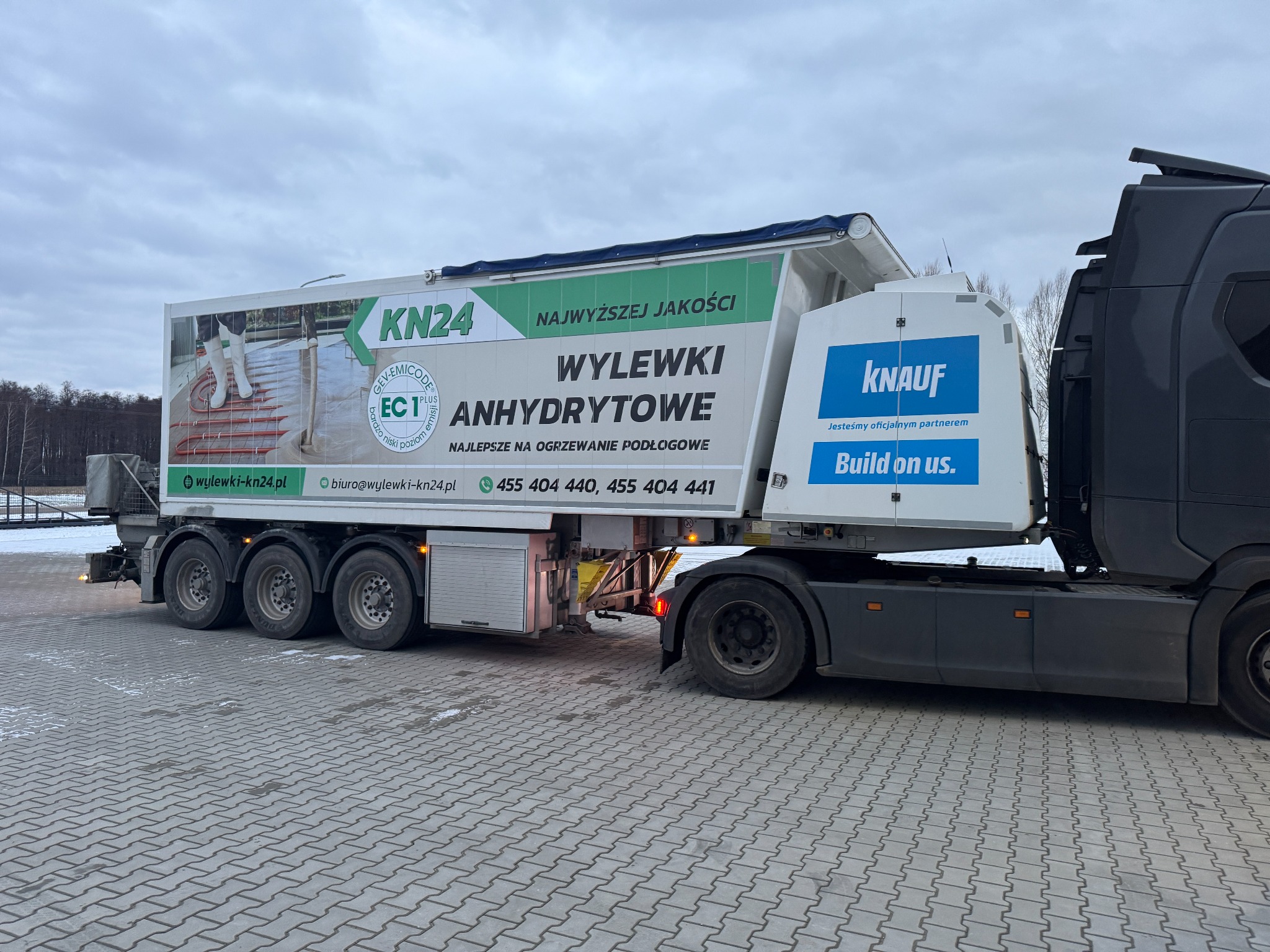 Ciężarówka z reklamą wylewek, zaparkowana w Wandowie na szarej kostce brukowej. Na naczepie widoczne logo KN24 i Knauf oraz hasła reklamowe.