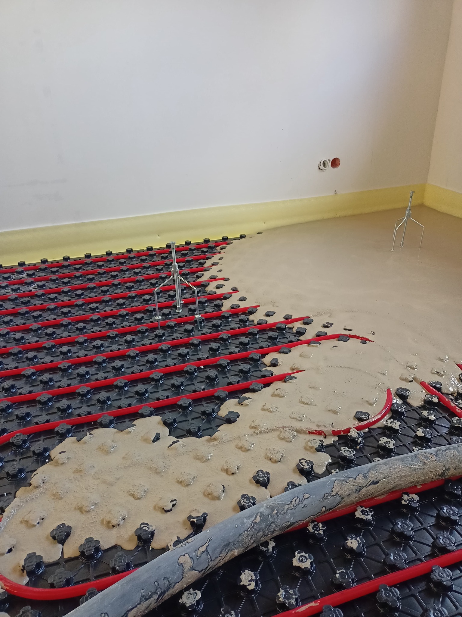 Rozlana masa na ogrzewaniu podłogowym z rurkami w kolorze czerwonym, częściowo zakrywająca czarną matę izolacyjną z wypustkami, widoczne metalowe stojaki poziomujące w rogu pomieszczenia.