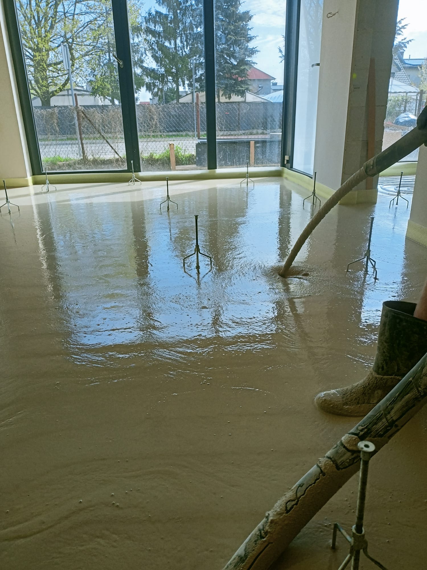 Wylewanie płynnej posadzki na podłogę z użyciem pompy; widoczne pionowe znaczniki wysokości i refleksy światła w świeżej wylewce, zarys buta roboczego.