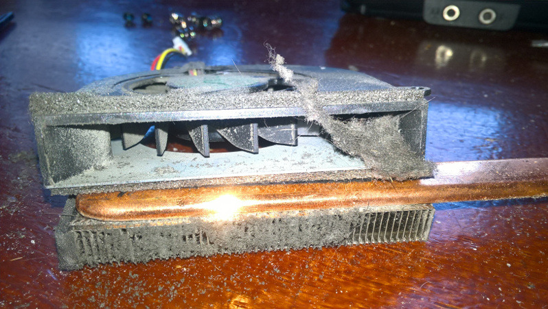 Zdemontowany, zakurzony radiator z wentylatorem laptopa, widoczne żebra chłodzące i miedziana rurka cieplna, drobne elementy elektroniczne w tle.