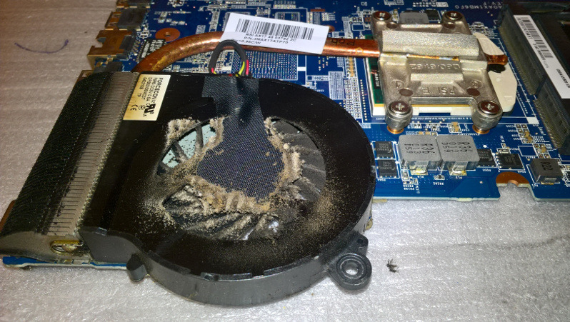 Zdemontowany, zakurzony wentylator chłodzenia laptopa, leżący obok płyty głównej. Widoczne żebra radiatora i miedziana rurka cieplna.