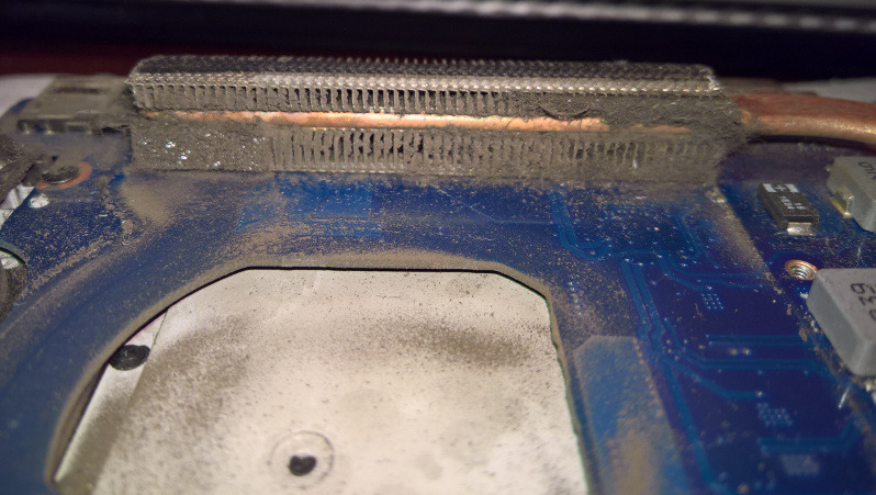 Zanieczyszczony radiator procesora w laptopie, pokryty grubą warstwą kurzu blokującą wylot powietrza, widoczna miedziana rurka cieplna i fragment płyty głównej.