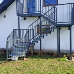 Zewnętrzne schody metalowe z balustradą prowadzące do drzwi na piętrze domu, widoczne trawnik i elewacja. Stalowa konstrukcja malowana na szaro.