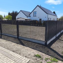 Stalsoft Mariusz Waśków - Nowoczesne, antracytowe ogrodzenie metalowe z pionowymi sztachetami, osadzone na betonowej podmurówce. W tle biały dom z ciemną dachówką i kościelna wieża.