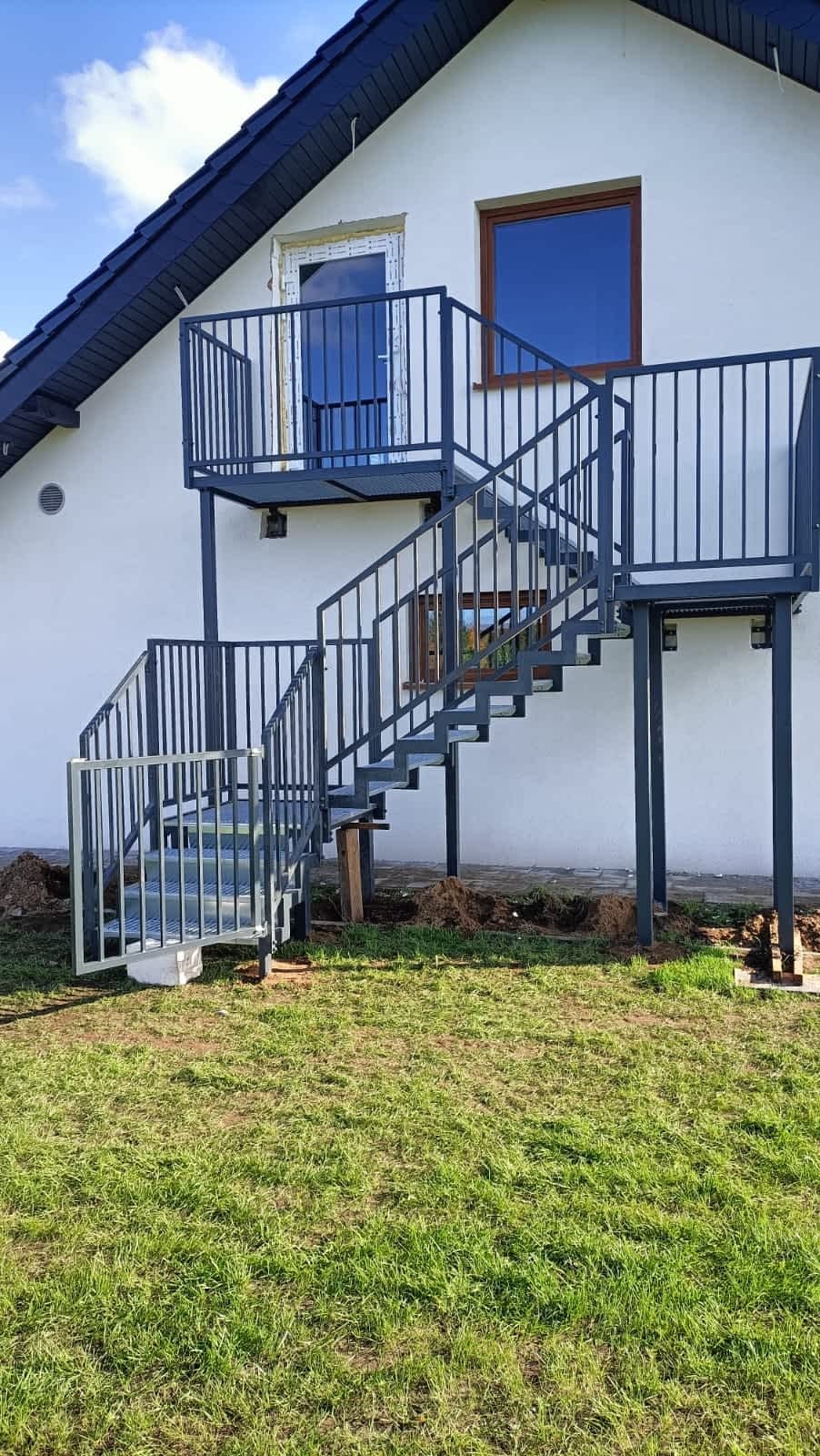 Zewnętrzne schody metalowe z balustradą prowadzące do drzwi na piętrze domu, widoczne trawnik i elewacja. Stalowa konstrukcja malowana na szaro.