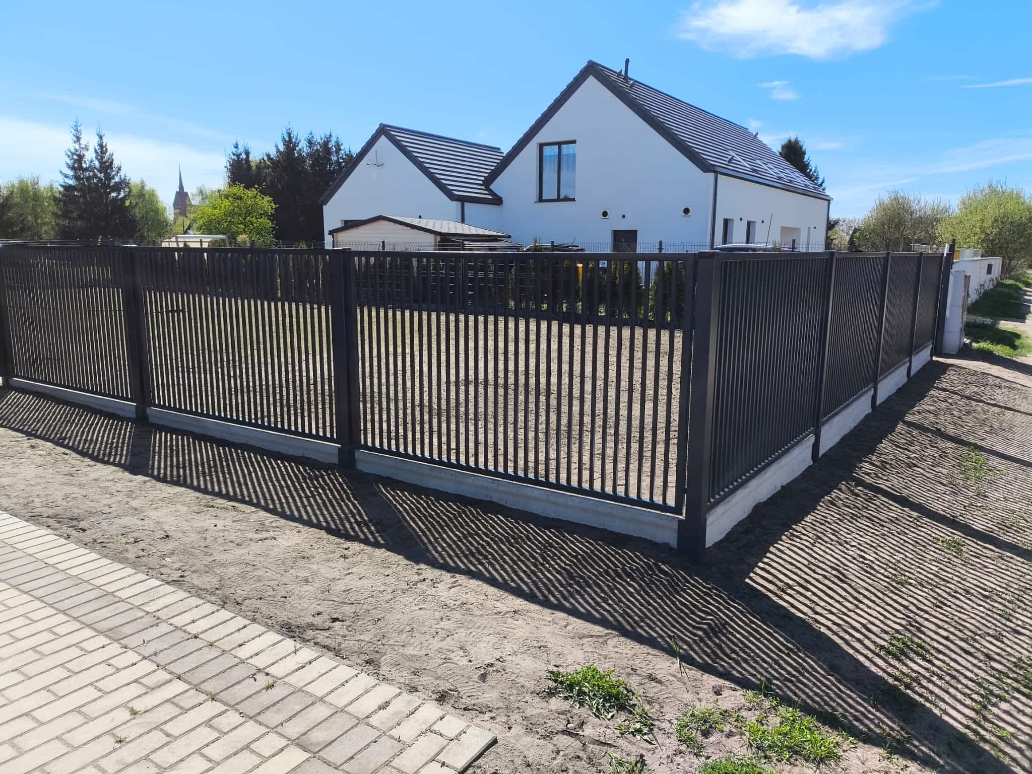 Nowoczesne, antracytowe ogrodzenie metalowe z pionowymi sztachetami, osadzone na betonowej podmurówce. W tle biały dom z ciemną dachówką i kościelna wieża.
