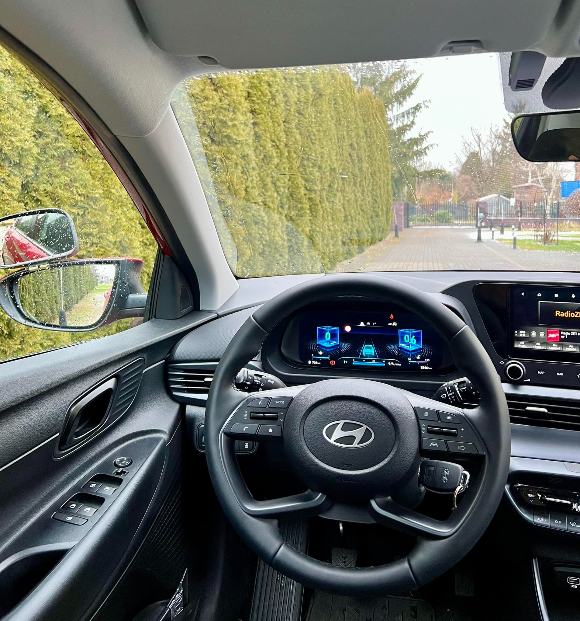 Wnętrze samochodu Hyundai z widocznym kokpitem, kierownicą z logo, cyfrowym wyświetlaczem i kluczykiem zwisającym z kierownicy, widok na drogę i zieleń za oknem.