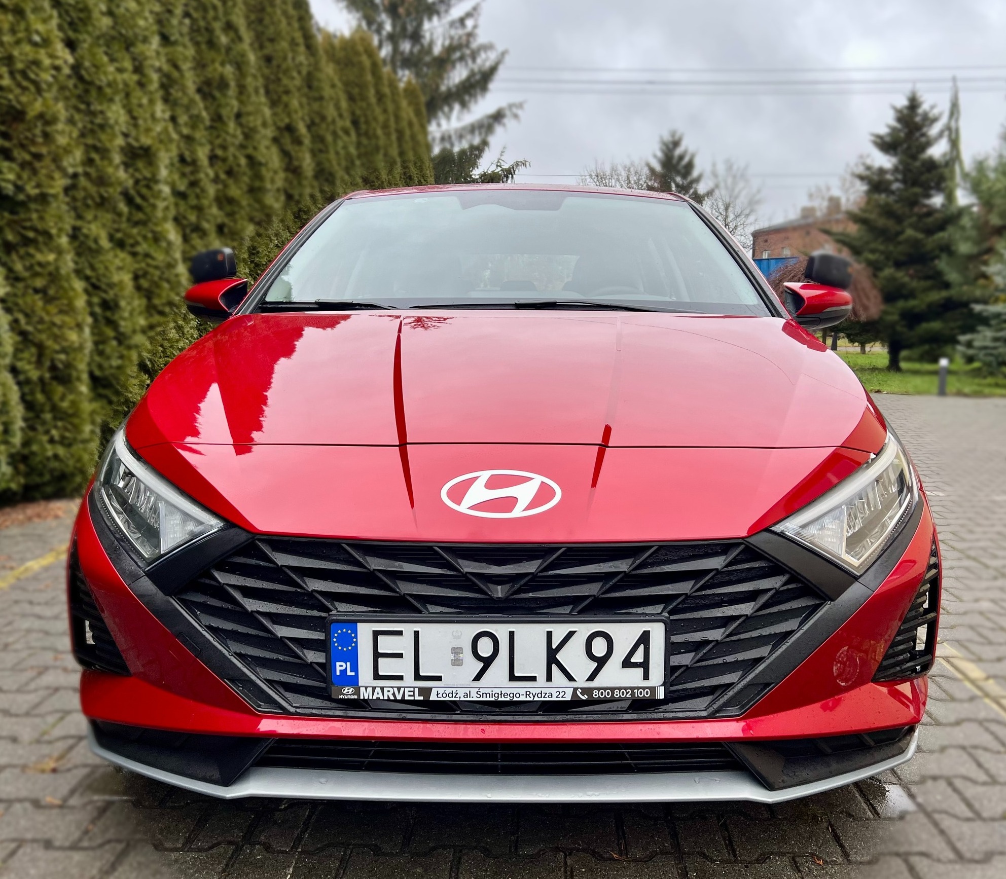 Przód czerwonego samochodu Hyundai z widoczną tablicą rejestracyjną EL 9LK94, logo marki i grillem, na tle zielonych krzewów i kostki brukowej.