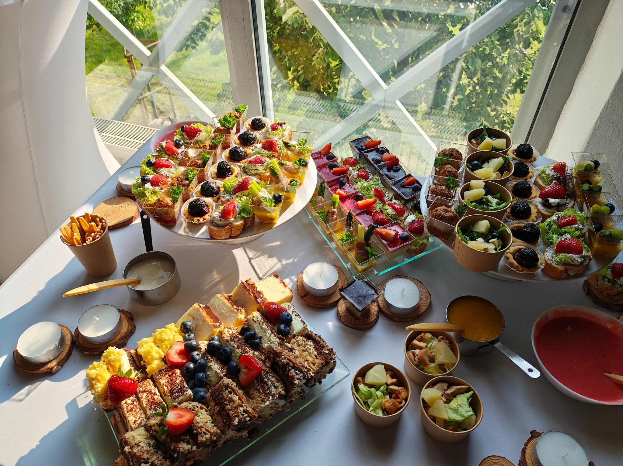 Dekoracyjny stół cateringowy z różnorodnymi przekąskami, ciastami i deserami, udekorowany świecami, z widokiem na zieleń za oknem.