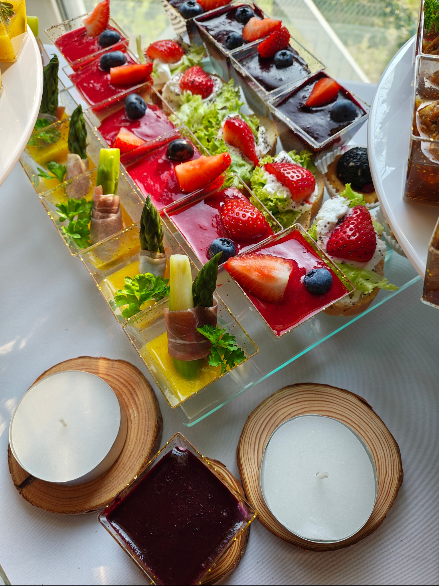 Elegancki catering: mini desery i przekąski w szklanych naczyniach, udekorowane owocami i ziołami, ustawione na stole z dekoracyjnymi świecami.