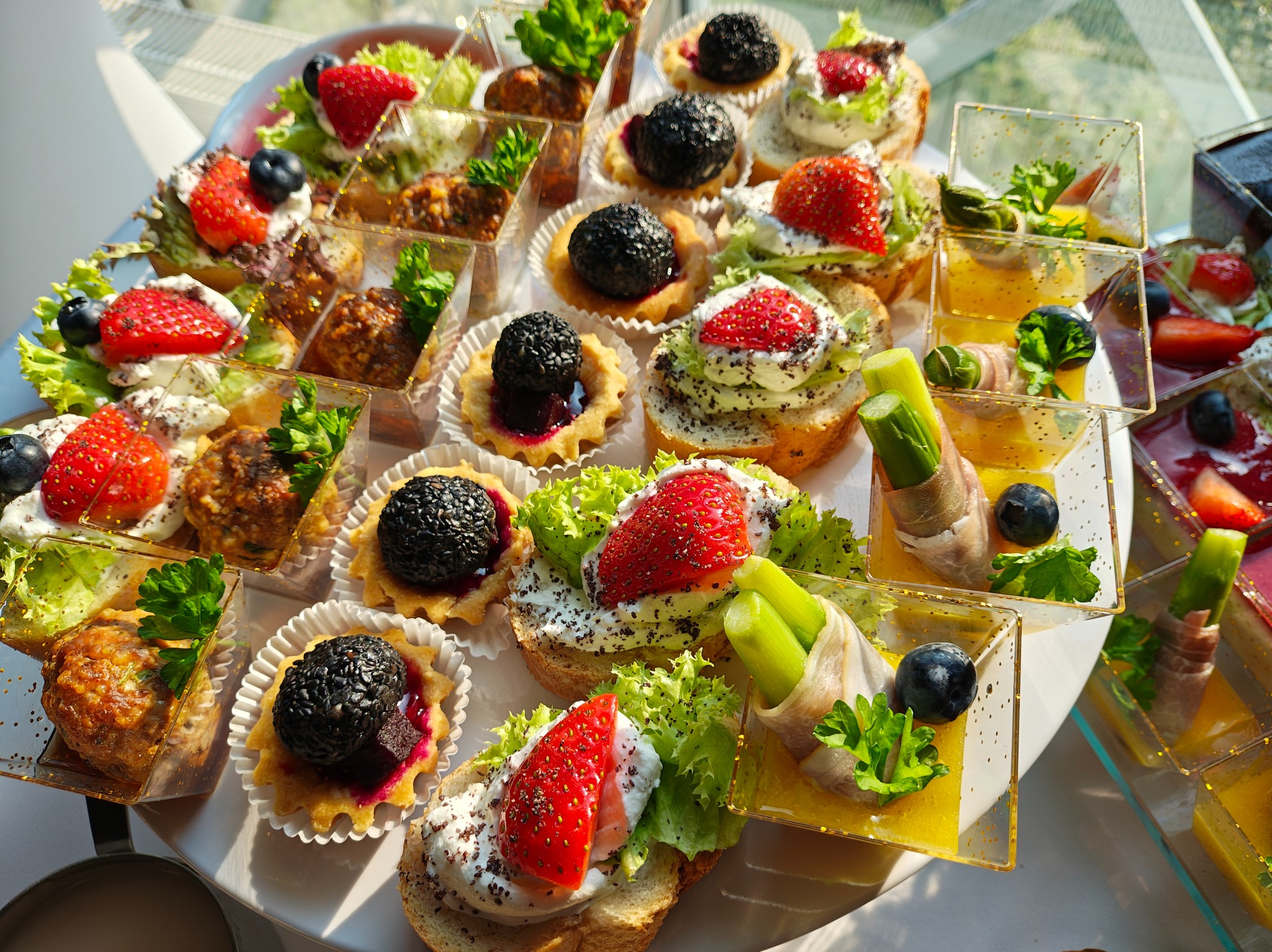 Elegancki catering: Różnorodne przekąski, w tym tartaletki z jeżynami, kanapki z truskawkami i sałatą oraz mięsne kulki w przezroczystych pojemnikach.
