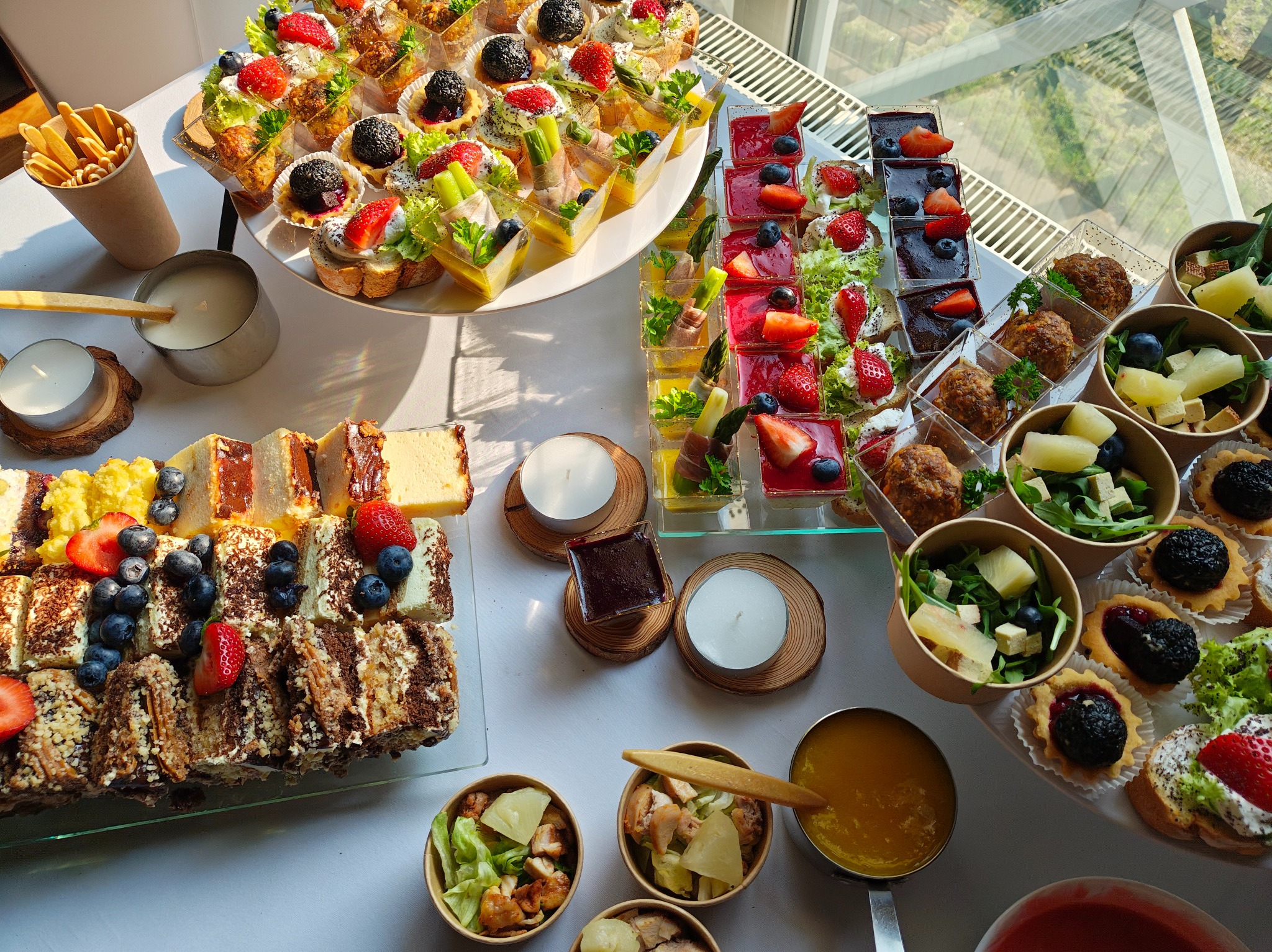 Bogato zastawiony stół z różnorodnymi przekąskami: tartaletki, mini-kanapeczki, sałatki w kubeczkach, ciasta z owocami, świece. Idealny catering na imprezę.