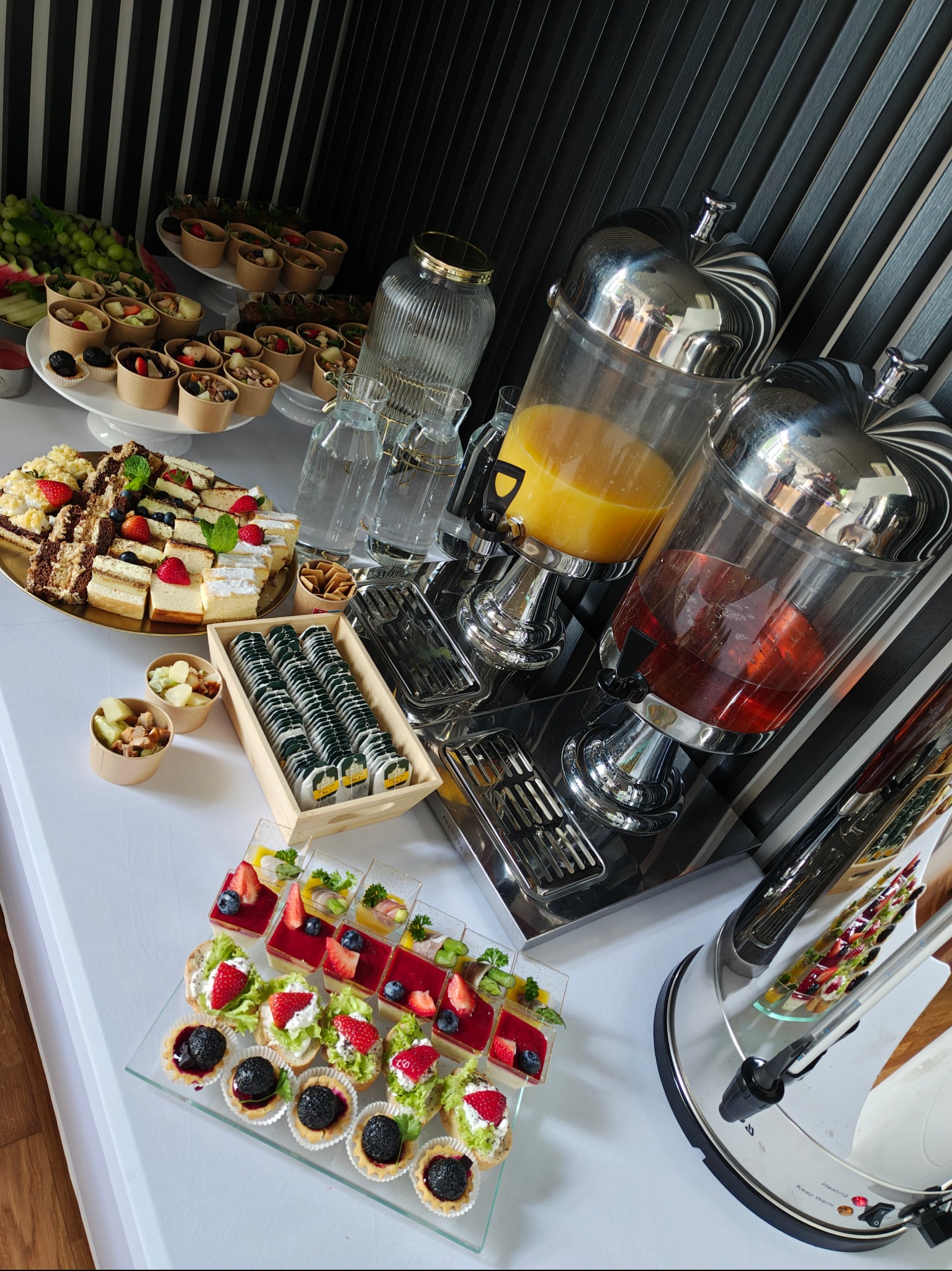 Stół cateringowy z napojami w dyspenserach i przekąskami: mini tartaletki, ciasta, sałatki w kubeczkach, owoce i herbata w torebkach, ustawione pod kątem.